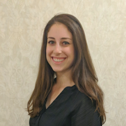 Woman with long brown hair wearing a black top against a beige wall.