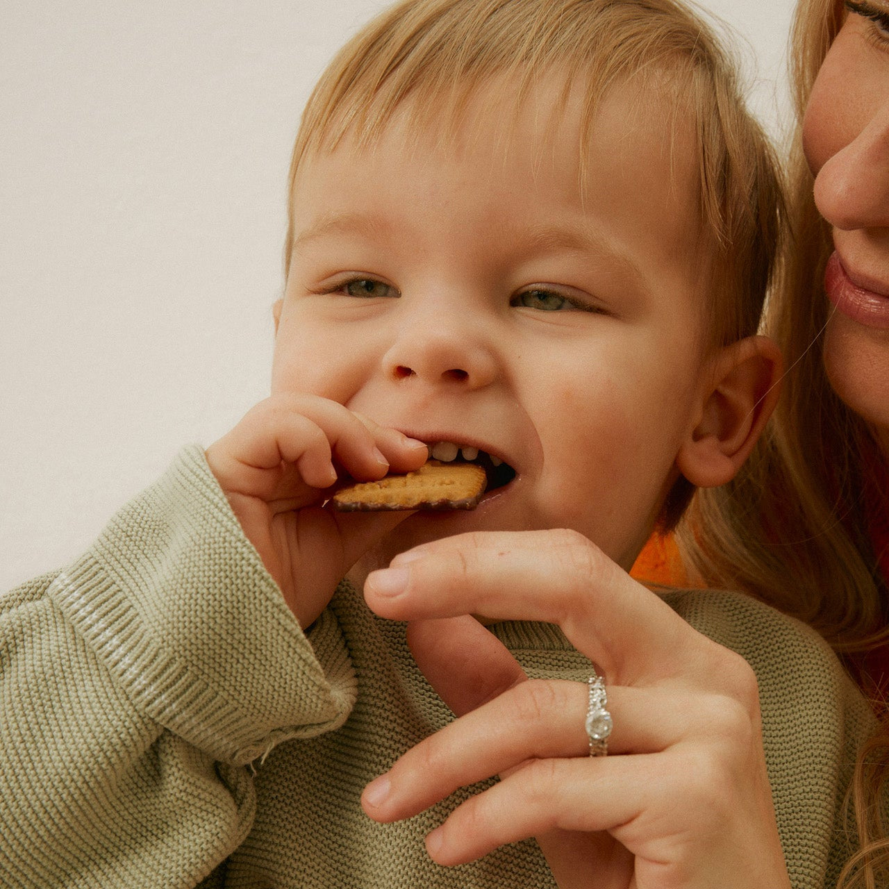 Child eating a cookie with a woman holding them, both wearing green sweaters.